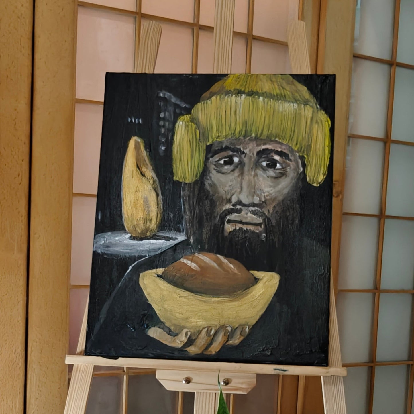 Painting of a face with a yellow hat on an easel in a room with plants and a lantern.
